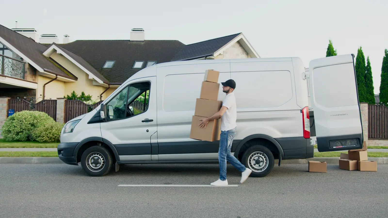Mann trägt gestapelte Pakete von einem weißen Transporter vor einem Wohnhaus, Symbolbild